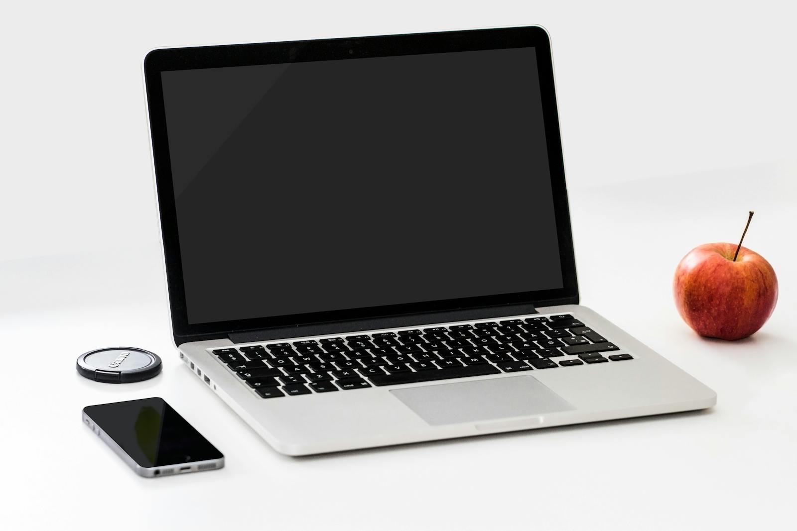 A minimalist workspace featuring a laptop, smartphone, and an apple on a clean white desk.